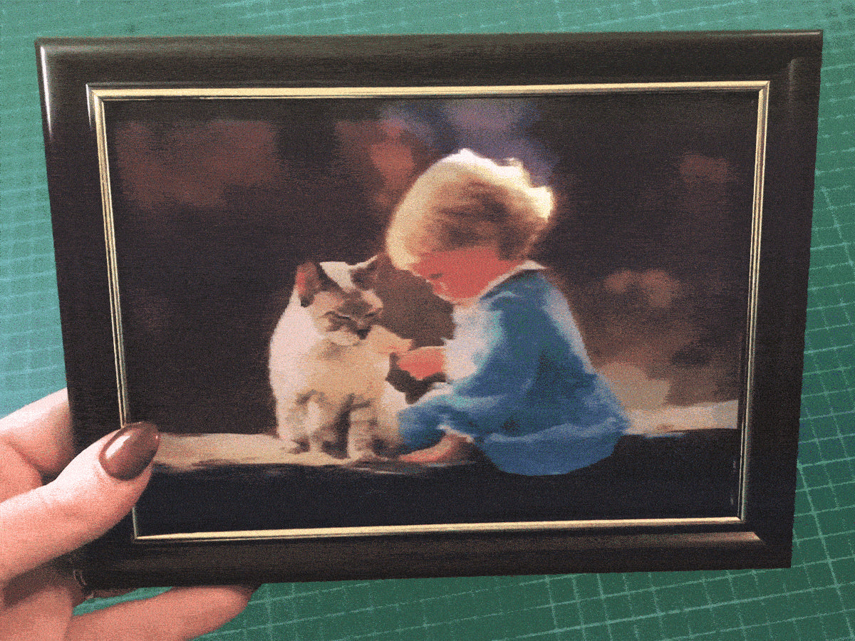 A framed lenticular image of a child in blue clothing petting a cat, held by a person with painted nails