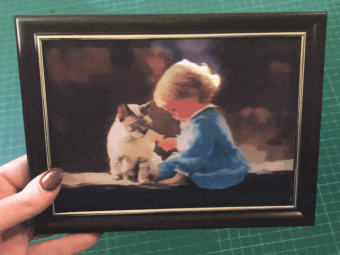 A framed lenticular image of a child in blue clothing petting a cat, held by a person with painted nails