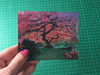 Hand holding a lenticular postcard showing a colorful tree with shifting shades and details.