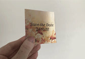 A person holds a beige lenticular 'Save the Date' card with floral designs. When tilted, the card transitions between two images—one displaying the text 'Save the Date' with the date '24/05/22,' and the other revealing additional decorative elements.